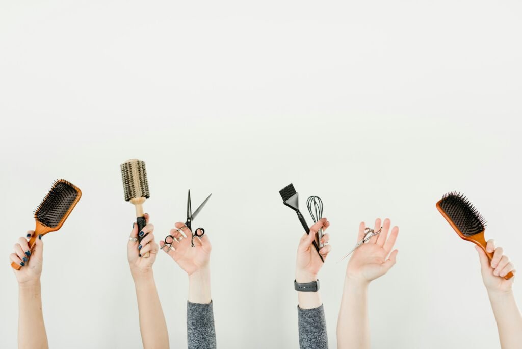 Hands raised holding assorted hair styling tools (brushes, scissors, curling irons, combs) against a pale background, showcasing a hairdressing theme.