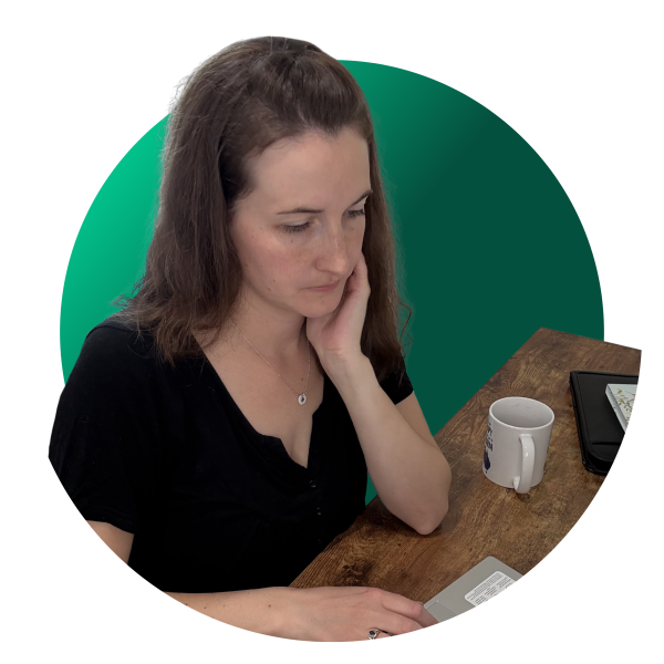 Woman with brown hair sits at a wooden desk, resting her chin on her hand and looking down at a laptop.